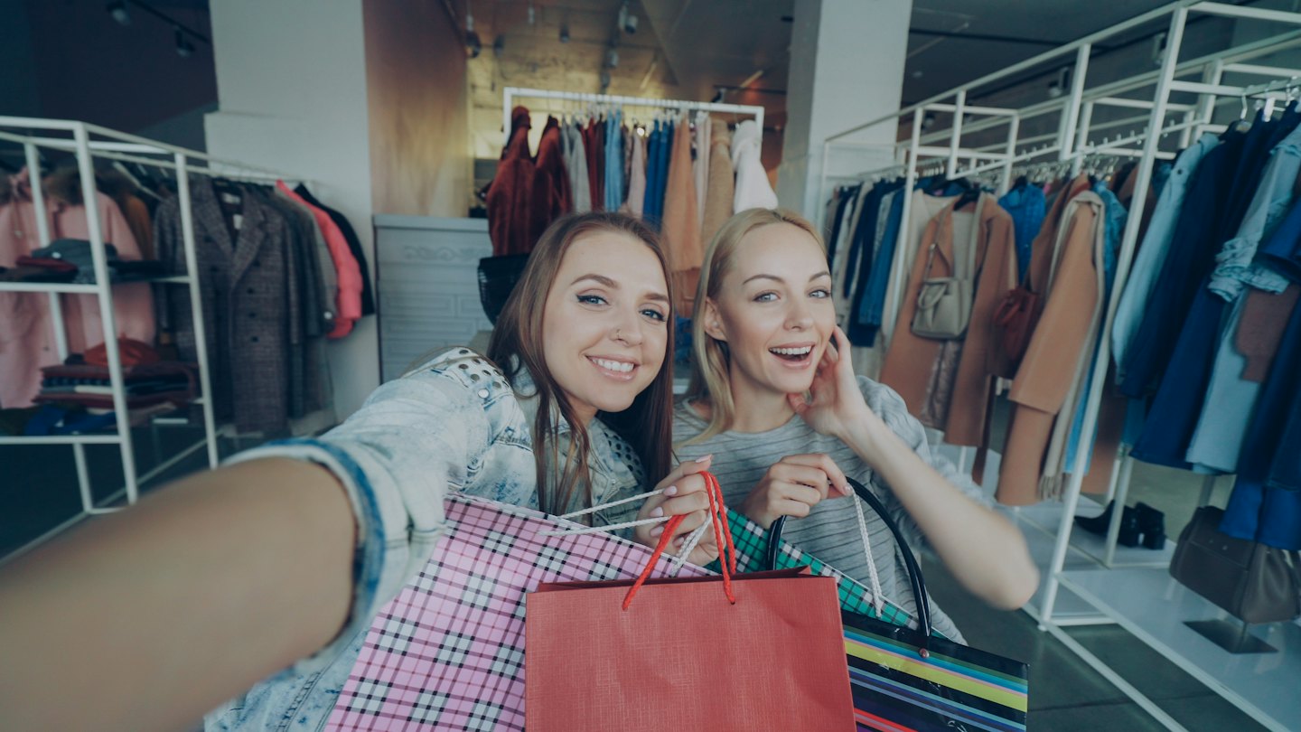 Two women take a selfie with shopping bags in a boutique.